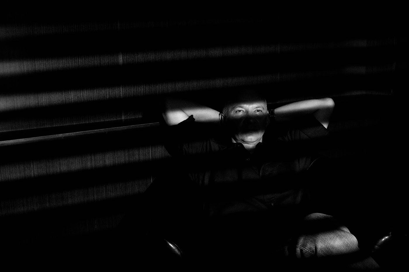 Black-and-white portrait of Artur Ekert leaning back with hands behind his head, his face crossed by bands of light through window blinds